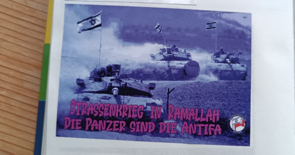 A photograph of three modern tanks waving Israeli flags while driving over barren hills, tinted dark blue. “Street war in Ramallah / The tanks are the antifa.”
