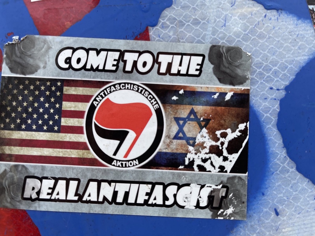 The US flag appears on the left, the Israeli flag on the right, both treated with a grimy filter. Connecting the two of them is an Antifascist Action logo, its flags red and black. “Come to the real antifascist” (sic)