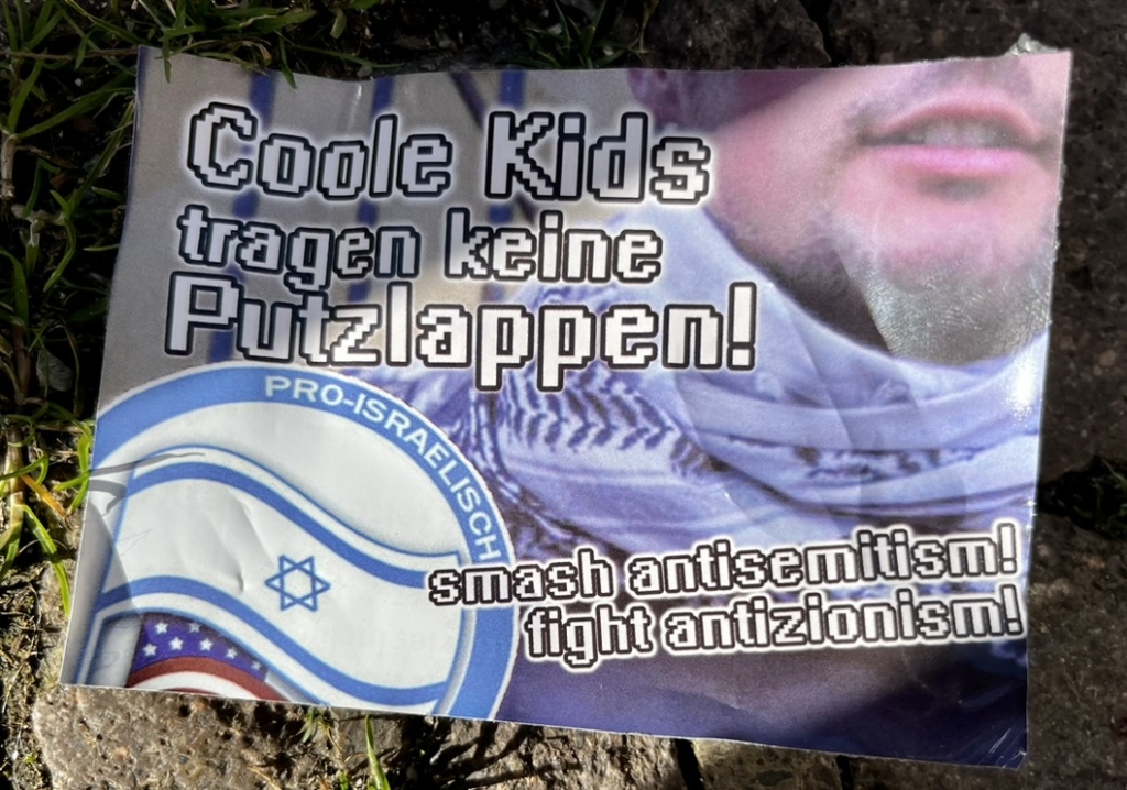 The shoulder, mouth, and jaw of a white man wearing a black-and-white kaffiyeh. “Cool kids don’t wear cleaning rags!” The lower-left corner features a take on the Antifascist Action logo featuring the text “Pro-Israelisch” with an Israeli flag with a US flag visible in the background. “Smash antisemitism! Fight antizionism!”