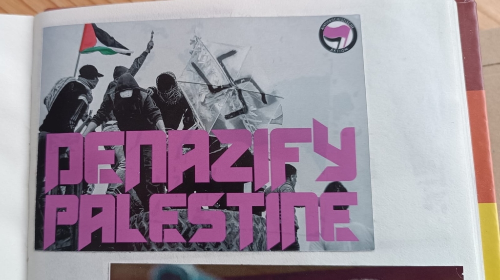 A photo of Palestinian protestors waving a Palestine flag and holding a kite adorned with a graffiti swastika. “Denazify Palestine.”