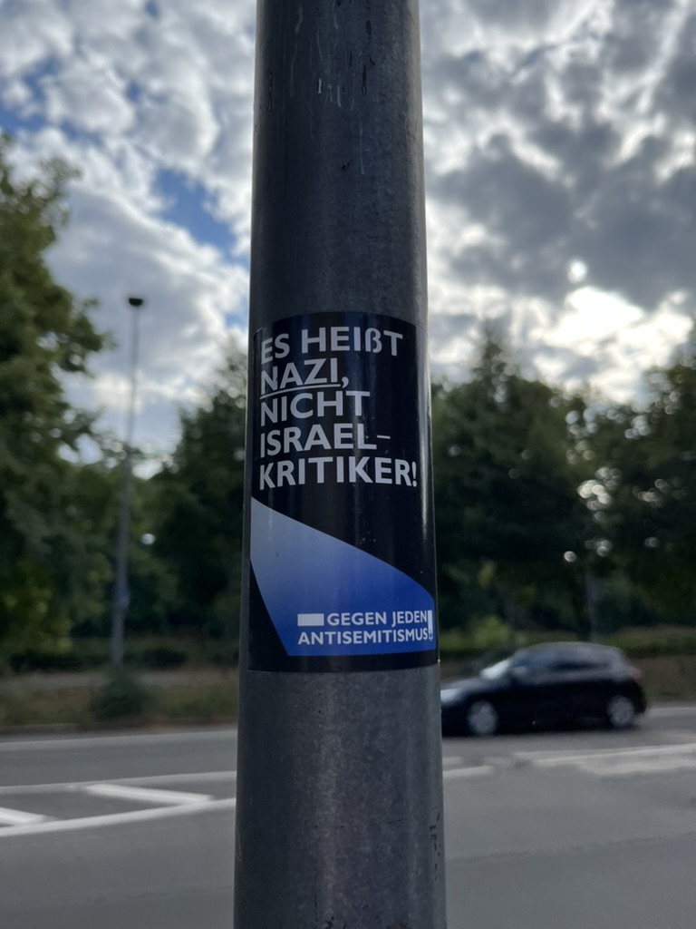 On a lamppost, a vertical sticker reads “It’s called NAZI, not Israel-critic!” In the path graphic below, “Against any and every antisemitism.”