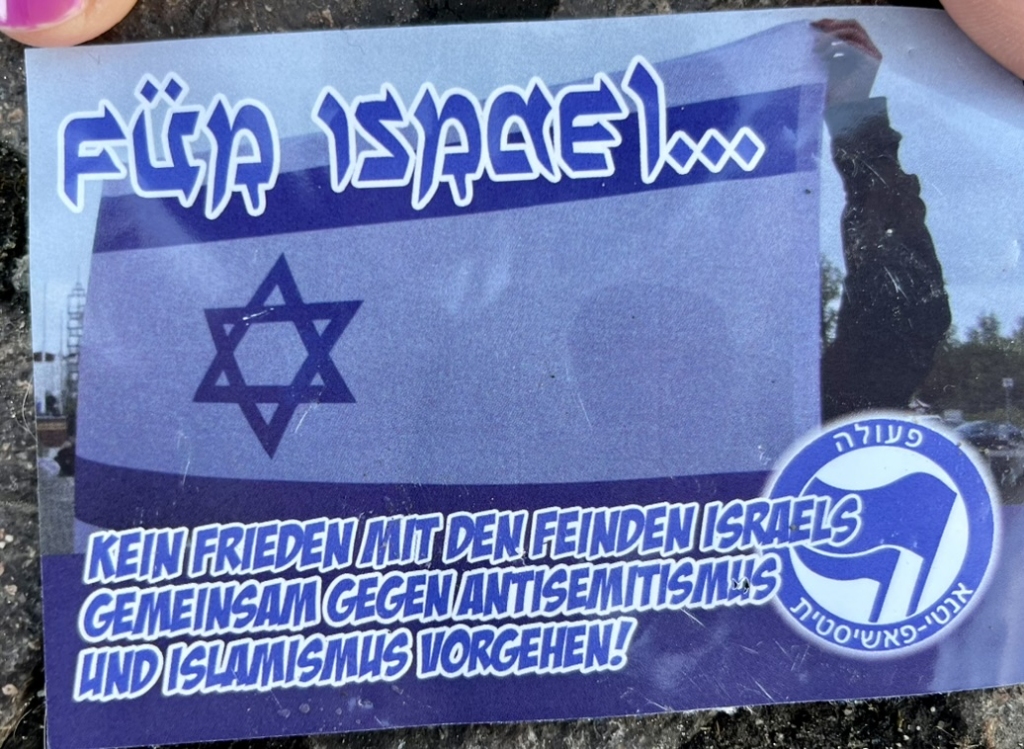 A photo of an Israeli flag. Across the top of the photo, a pseudo-Hebrew font set in mid blue with a white outline, reads “For Israel…”. Across the bottom of the sticker: “no peace with the enemies of Israel / Act together against antisemitism and Islamism!”