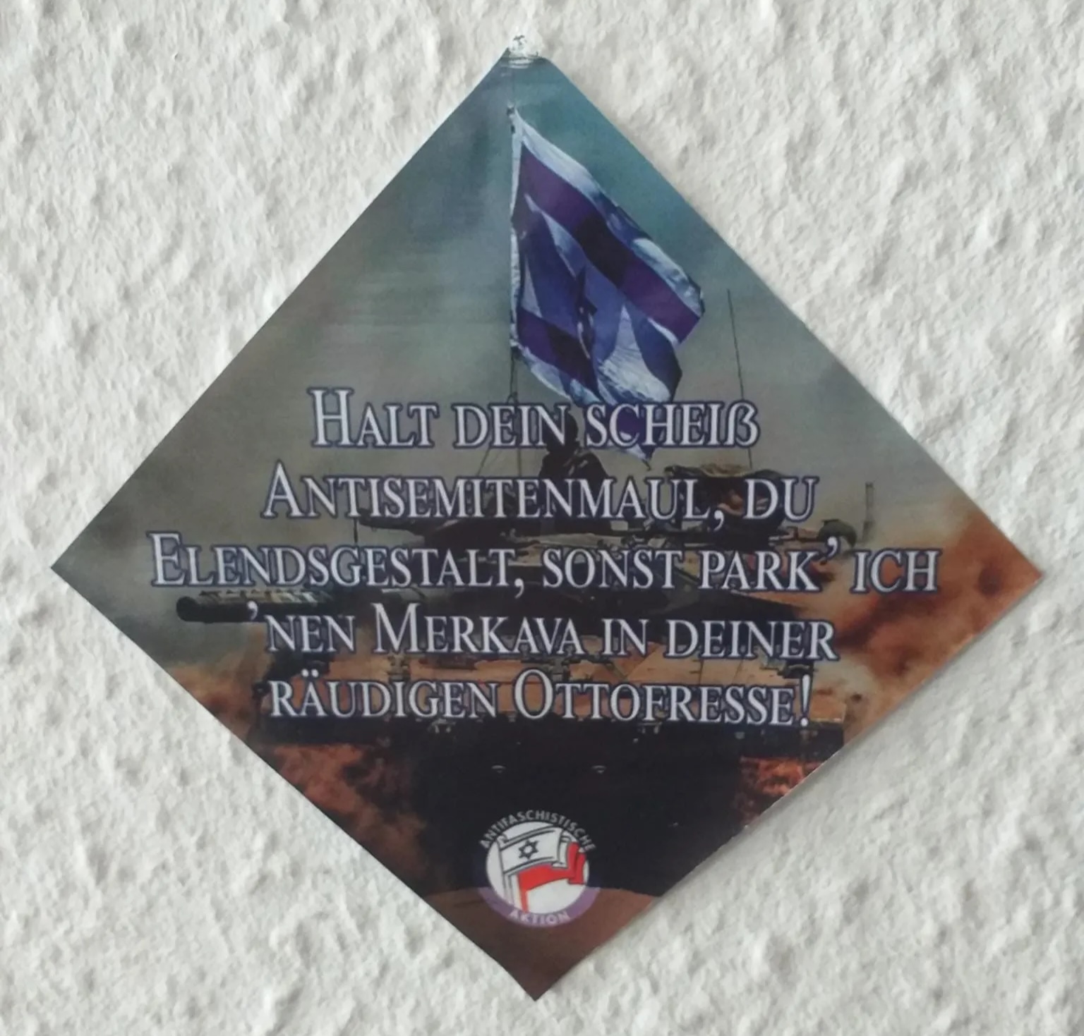 A square sticker showing a full-color photograph of a modern tank with a massive Israeli flag waving above it. “Shut your scheiß antisemite mouth, you miserable wretch, or I’ll park a Merkava in your dirty fool face.”