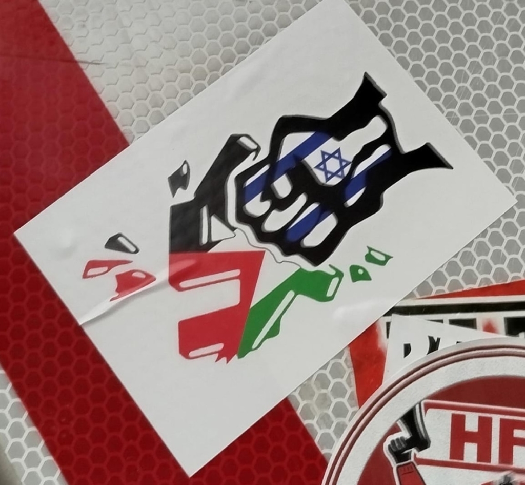 A stylized fist smashing a swastika. The fist is overlaid with the flag of Israel, while the swastika and the splinters coming off of it are overlaid with the Palestinian flag.