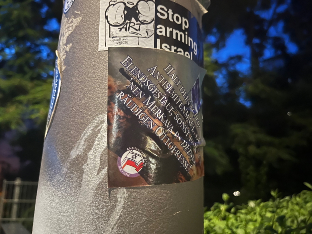 On a lamppost, a small horizontal black sticker states simply “Stop arming Israel”, in English. Covering its very bottom is the square sticker described earlier in this article, showing an Israeli tank and German text reading “Shut your scheiß antisemite mouth, you miserable wretch, or I’ll park a Merkava in your dirty fool face."