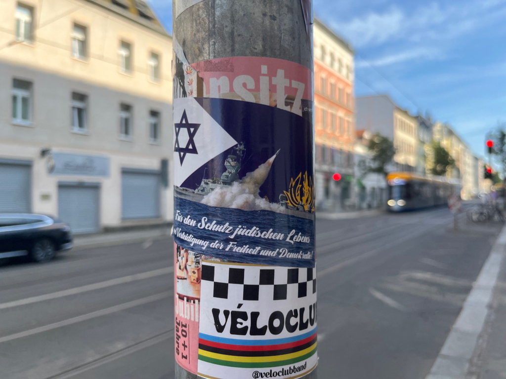 On a lamppost covered in other (non-political) stickers, a blue sticker displays a photo of a warship launching a rocket with a dark blue Star of David in the background. The emblem of the Israeli navy is stamped on the background in yellow. “For the protection of Jewish life and the defense of freedom and democracy!”